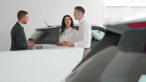 Gerente de ventas ayudando a la joven pareja a elegir un coche nuevo en la sala de exposición de coches modernos. Pareja joven hablando con un vendedor en una sala de exposición de coches. - Metraje, vídeo