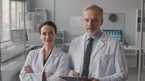 Średni portret pewnej siebie kaukaskiej pary ekspertów medycznych ubranych w białe fartuchy laboratoryjne pozujących do kamery w przestronnej nowoczesnej klinice - Materiał filmowy, wideo