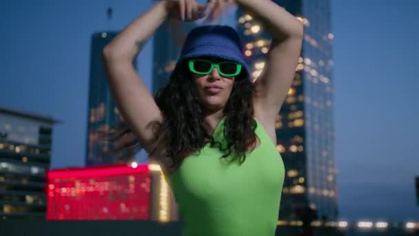 Активна афроамериканська чорна жінка в неоновому зеленому верху і фіолетовий відро капелюх танцює на фоні міських вогнів у центрі міста вночі. Прохолодна талановита змішана гонка жіноча танцівниця рухається ритмічно 4K Slowmo - Кадри, відео