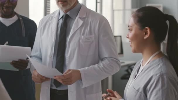 Średnie ujęcie białego mężczyzny w średnim wieku głównego lekarza mówiącego do wielorasowych kolegów stojących razem w jaskrawo nowoczesnym gabinecie lekarskim - Materiał filmowy, wideo