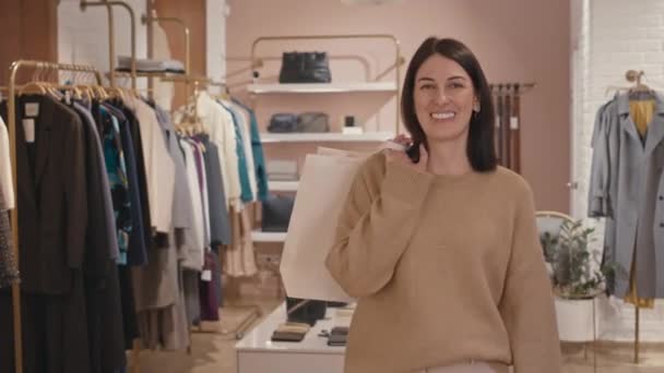 Medium portrait of cheerful young Caucasian woman with white paper shopping bags posing for camera in contemporary clothes store - Footage, Video