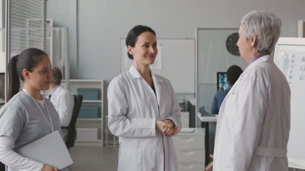 Inyección media de dos doctoras multiétnicas con batas blancas de laboratorio estrechándose la mano después de tener una conversación en el lugar de trabajo - Metraje, vídeo