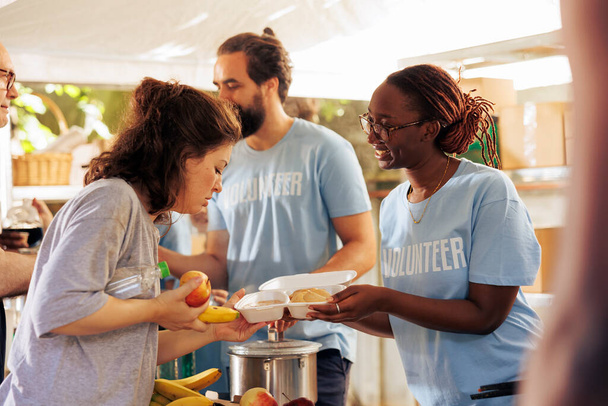 African american volunteer lady at local center actively involved in distributing nutritious meals to alleviate hunger among the less fortunate. Black woman helper sharing food to homeless person. - Photo, Image