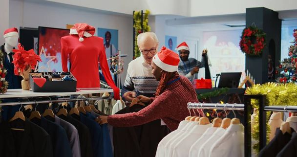 Ηλικιωμένος πελάτης που βοηθιέται από το λιανικό βοηθό στο εορταστικό διακοσμημένο κατάστημα ρούχων για να καθορίσει εάν το κομψό σακάκι είναι η σωστή εφαρμογή. Υπάλληλος που βοηθά ηλικιωμένο άνδρα στο χριστουγεννιάτικο στολισμένο κατάστημα μόδας - Φωτογραφία, εικόνα