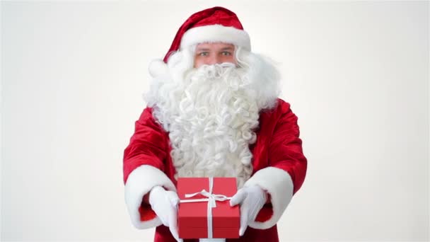 サンタクロースは彼の手に贈り物を持って提供しています。白い背景に幸せなサンタを微笑む。面白いサンタクロースはスタジオでポーズをとっています。居心地の良いクリスマスと新年2016の概念。クリスマスプレゼント - 映像、動画