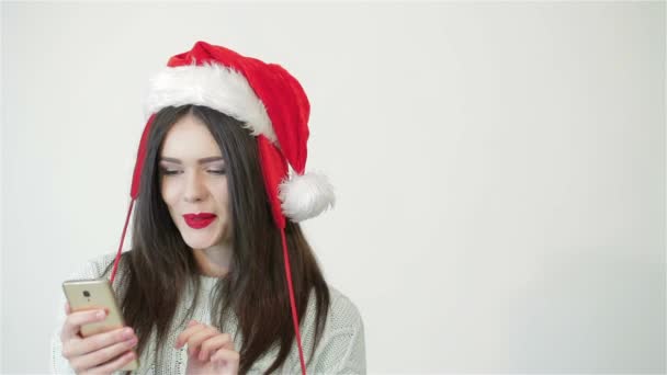 Дівчина в капелюсі Санта використовує смартфон і виглядає побічно копіюючи простір. Посміхаючись щасливій дівчині на білому тлі. Красива жінка, дівчина, жінка з капелюхом Крістенмаса позує в студії. різдвяні подарунки - Кадри, відео
