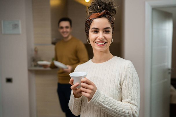 Одна молодая белая женщина, стоящая в своей квартире с чашкой кофе, в то время как ее муж или парень находится на кухне ежедневно утром пара рутинная счастливая улыбка реальные люди копируют пространство - Фото, изображение