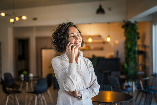 One woman mature caucasian female businesswoman entrepreneur stand at work or home use mobile phone making a call talk real people copy space wear white shirt curly hair happy smile - Foto, imagen