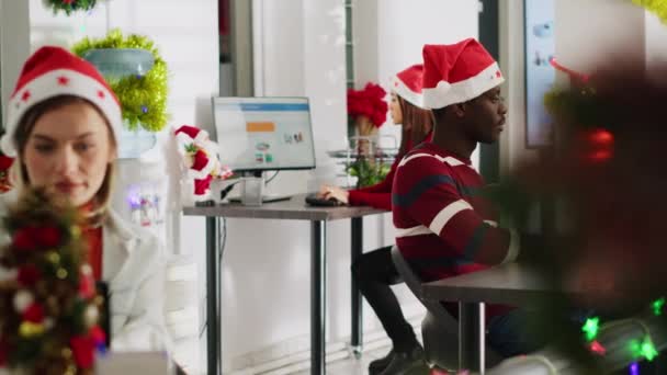 Portrait of friendly african american startup employee working on christmas season in decorated office. Workers solving tasks at workstation in multiethnic workplace during winter festive time - Footage, Video
