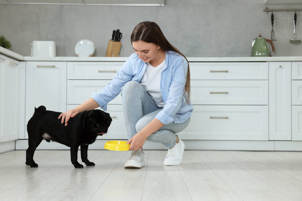 Beautiful young woman feeding her adorable Pug dog in kitchen - Zdjęcie, obraz