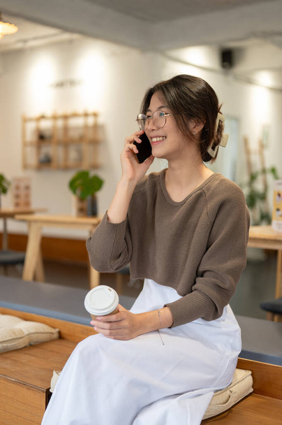 Μια όμορφη και χαρούμενη νεαρή Ασιάτισσα απολαμβάνει να μιλάει στο τηλέφωνο με τη φίλη της ενώ χαλαρώνει σε μια όμορφη, ζεστή καφετέρια. Έννοια τρόπου ζωής - Φωτογραφία, εικόνα