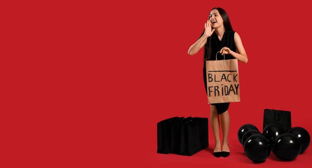 テキスト用のスペースを持つ赤い背景のショッピングバッグと風船を持つ美しい若い女性. ブラックフライデーセール - 写真・画像