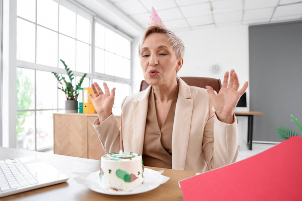 Mujer madura con pastel de cumpleaños haciendo deseo en la oficina - Foto, Imagen