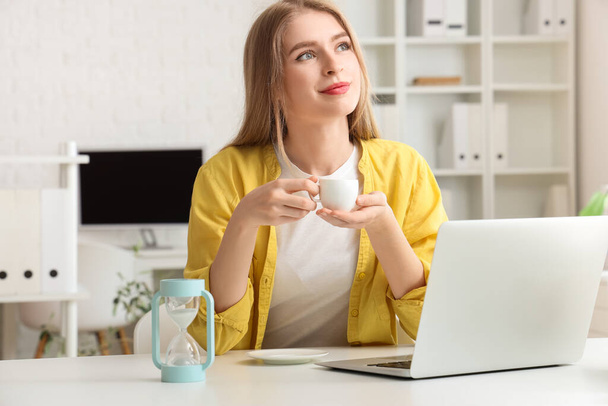 Молодая деловая женщина с чашкой кофе и песочными часами в офисе - Фото, изображение