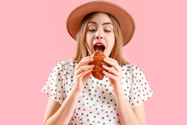 ピンクの背景でおいしいクロワッサンを食べるウィッカー帽子の美しい若い女性 - 写真・画像