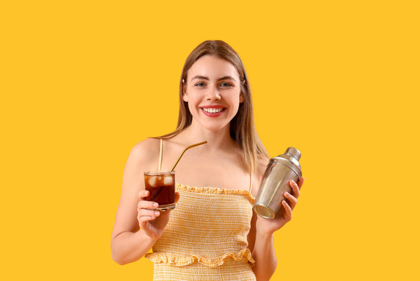 Beautiful happy young woman with glass of tasty cola and shaker on yellow background - Photo, Image