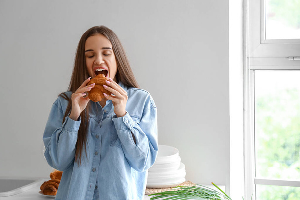 Beautiful young woman eating tasty croissant in kitchen - Photo, Image