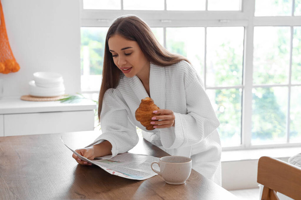 Hermosa mujer joven en albornoz con sabroso croissant y taza de café periódico de lectura en la cocina - Foto, imagen