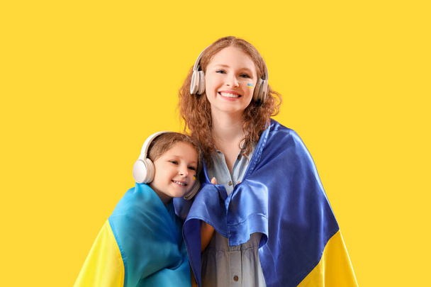 Glückliches kleines Mädchen und ihre Mutter mit ukrainischen Flaggen, die Musik über Kopfhörer auf gelbem Hintergrund hören - Foto, Bild