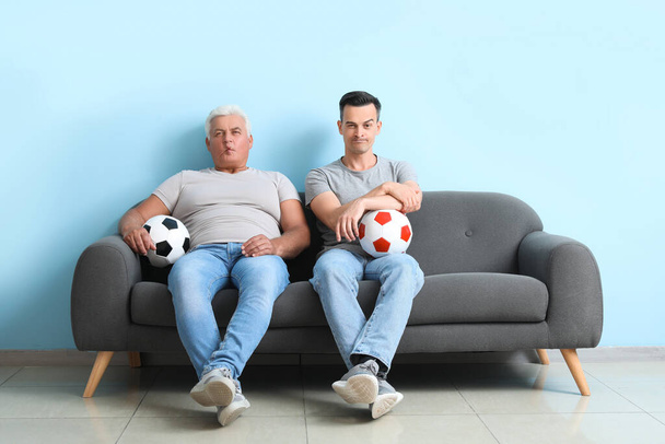 Молодой человек со своим отцом и футбольными мячами сидит на диване рядом с синей стеной - Фото, изображение