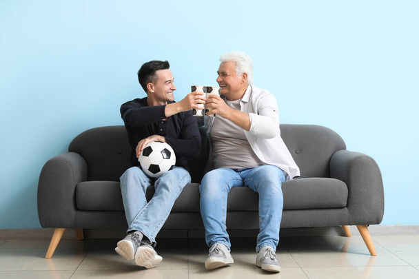 Egy fiatalember az apjával focimeccset néz a kanapén a kék fal mellett. - Fotó, kép