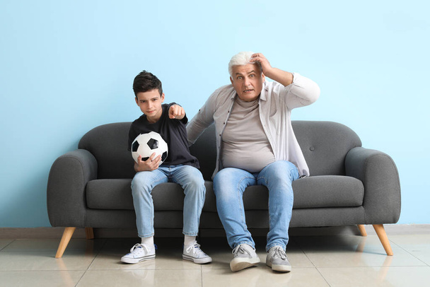 Мальчик с дедушкой смотрят футбол на диване возле голубой стены. - Фото, изображение