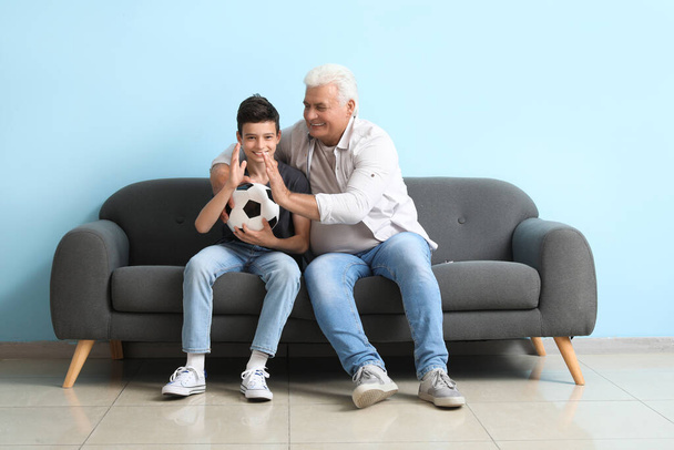 Mały chłopiec z dziadkiem ogląda mecz piłki nożnej na kanapie w pobliżu niebieskiej ściany - Zdjęcie, obraz