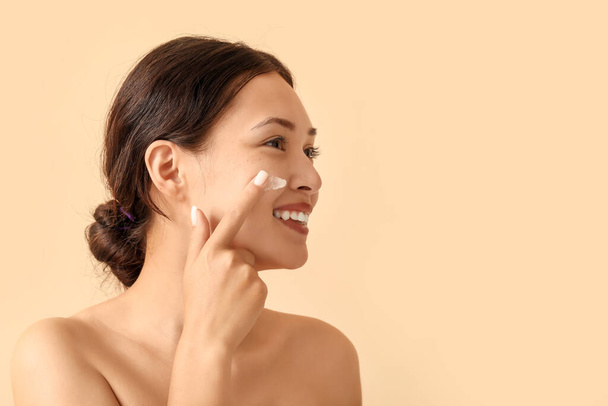 Beautiful Asian woman applying facial cream on beige background, closeup - Photo, Image