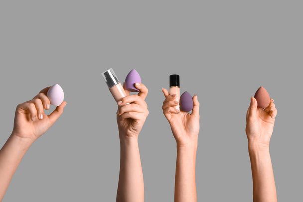 Жіночі руки з губками для макіяжу та пляшками фундаменту на сірому фоні - Фото, зображення