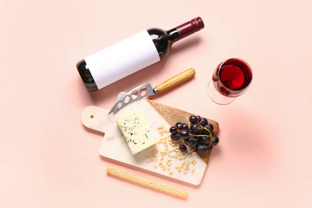 Board with tasty cheese, nuts, grapes, bottle and glass of wine on pink background - Photo, Image
