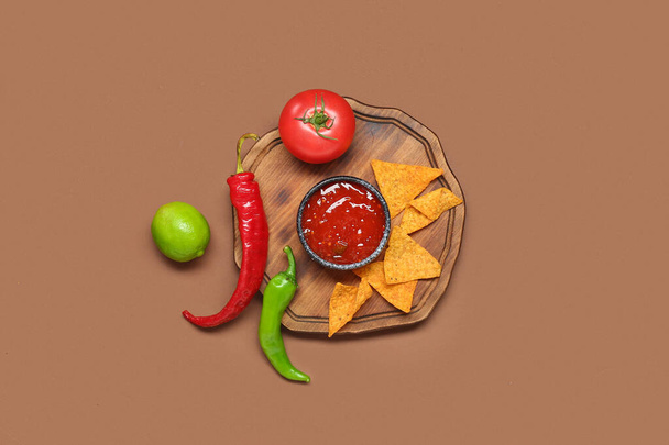 Wooden board with tasty salsa sauce, nachos and ingredients on brown background - Photo, Image