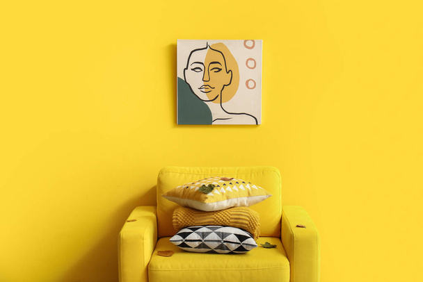 Sessel mit Stapel weicher Kissen und Malerei in der Nähe der gelben Wand - Foto, Bild