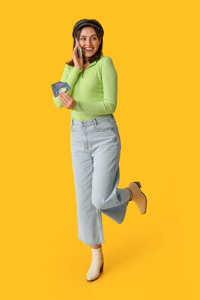黄色の背景で携帯電話で話すクレジットカードを持つ若い女性 - 写真・画像