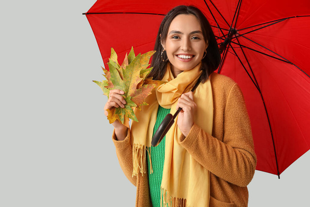 Beautiful young woman with autumn leaves and umbrella on grey background - Photo, Image