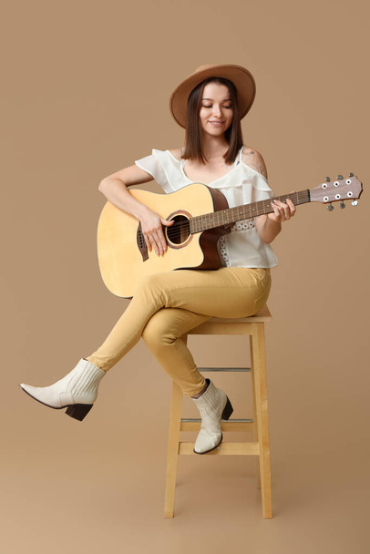 Mulher bonita tocando guitarra acústica enquanto sentada na cadeira contra fundo bege - Foto, Imagem