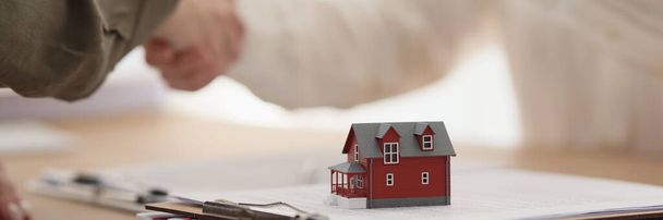 Immobilienmaklerin übergibt Schlüssel zu Haus an Klientin beim Händeschütteln. Kleines Modell eines Stadthauses steht auf einem Stapel Unterlagen auf dem Tisch im Büro - Foto, Bild