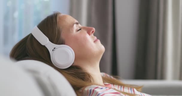 Fiatal nő pihentető kényelmes kanapé csukott szemmel visel fejhallgató, lány élvezi hallgatni a zenét és a hang pihentető otthon. Kiváló minőségű 4k felvételek - Felvétel, videó