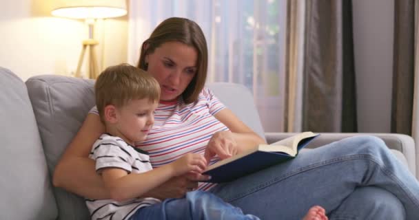 Anya és fia könyvet olvasnak egy kényelmes kanapén ülve a nappaliban, családi körben. Kiváló minőségű 4k felvételek - Felvétel, videó
