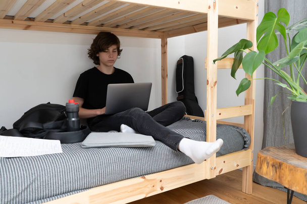 Nastoletni uczeń lub student siedzi na niższym poziomie łóżka piętrowego w swoim pokoju z laptopem w rękach i odrabia lekcje.. - Zdjęcie, obraz