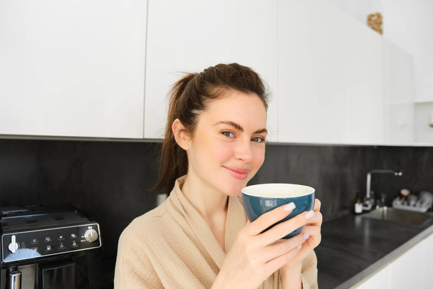 Ritratto di bella giovane donna in accappatoio, bere caffè del mattino e godersi il gusto, sorridente piacere, in piedi in cucina. - Foto, immagini