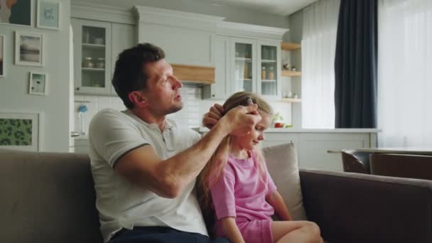 Opiekuńczy ojciec siedzi z córką w pokoju na kanapie, prostuje jej włosy i dekoruje je spinką do włosów. Rodzic i dziecko zrelaksować się po dniu roboczym po pracy, przedszkola lub szkoły w - Materiał filmowy, wideo