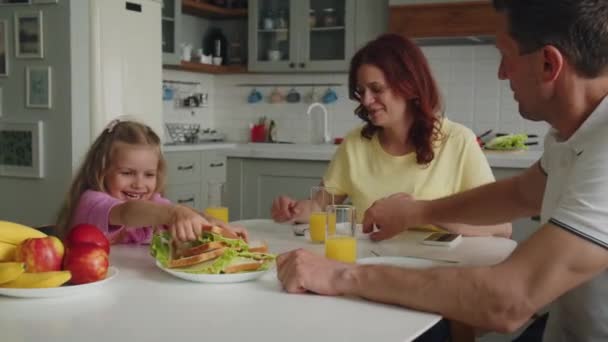 Une famille de rêve américaine souriante dans la cuisine prend le petit déjeuner ou le déjeuner. Fille met des sandwichs dans des assiettes pour la mère et le père. Maman la remercie chaleureusement, ainsi que son mari. Le concept de l'enfance heureuse et - Séquence, vidéo