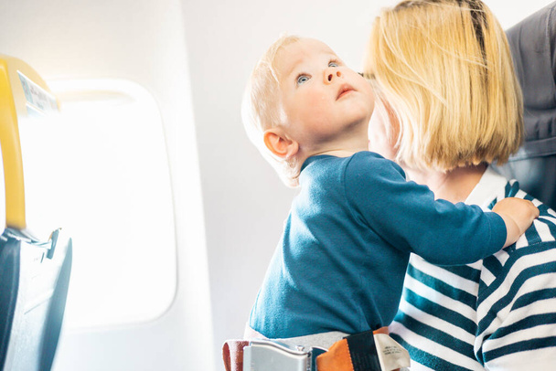 Η μαμά και το παιδί πετούν με αεροπλάνο. Μητέρα που κρατάει και παίζει με το βρέφος αγοράκι της στην αγκαλιά της κατά τη διάρκεια της οικονομικής πτήσης. Concept φωτογραφία του αεροπορικού ταξιδιού με το μωρό. Πραγματικοί άνθρωποι - Φωτογραφία, εικόνα