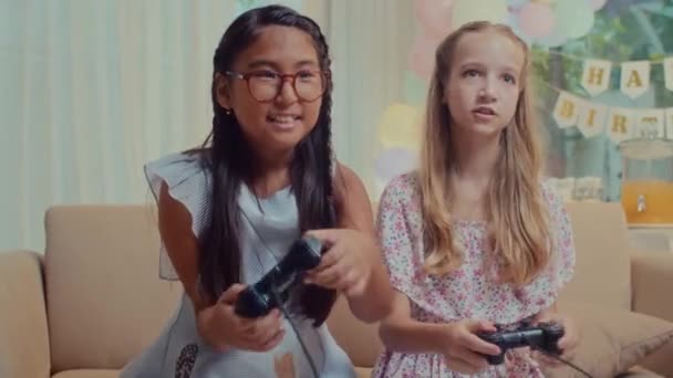 バックグラウンドで誕生日デコレーションで自宅でビデオゲームをするジョイスティックと幸せな女の子のミディアムショット - 映像、動画