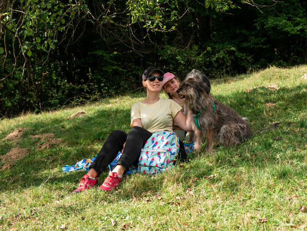 Gelukkig familie moeder met dochter en twee honden zitten op groen gras gazon plezier picknick tijd doorbrengen samen wandelen in de bergen. Mensen en huisdieren buiten levensstijl foto openhartige emoties. - Foto, afbeelding