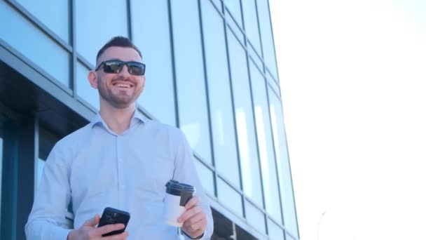 Sorridente uomo ricco che tiene smartphone e caffè in mano mentre in piedi vicino al suo centro ufficio. Gente felice. - Filmati, video