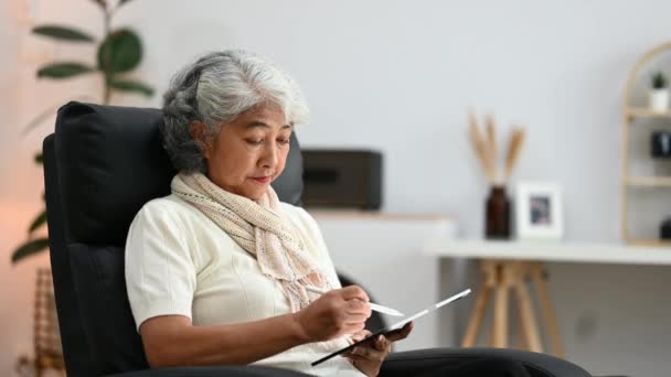Mujer de pelo gris de los años 70 feliz usando la tableta digital mientras que se sienta en el sillón en el país. - Metraje, vídeo