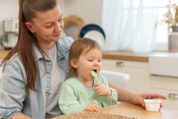 Happy family at home. Mother feeding her baby girl from spoon in kitchen. Little toddler child with messy funny face eats healthy food at home. Young woman mom giving food to kid daughter - Photo, Image