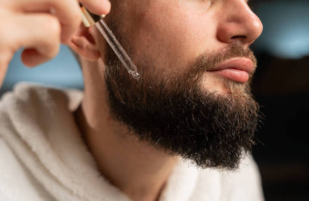 Primer plano de un hombre que se aplica aceite de barba en la cara en casa después de una ducha. Copiar espacio - Foto, imagen