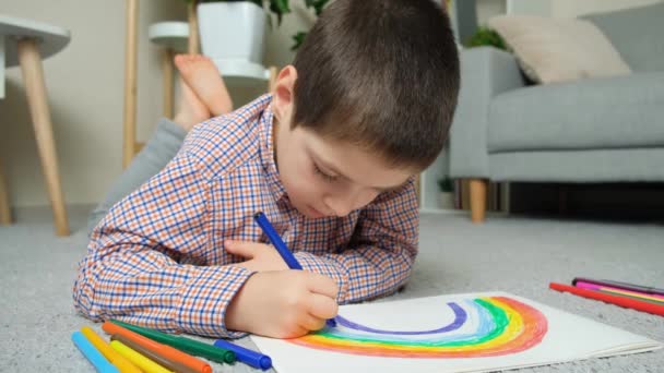 Ein kleiner Junge zeichnet einen Regenbogen mit Filzstiften in ein Skizzenbuch, während er zu Hause auf dem Teppich liegt. - Filmmaterial, Video